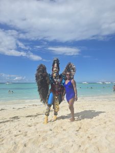 Two people pose on a sandy beach in Isla Mujeres - one in a blue swimsuit, the other in vibrant ceremonial attire with a feathered headdress, face paint, and bold costume—set against turquoise waters and anchored boats.